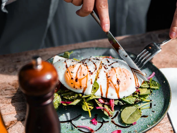 Breakfast at moxxa.caffè Hands are cutting fried eggs over a salad with balsamic vinegar on a wooden table.