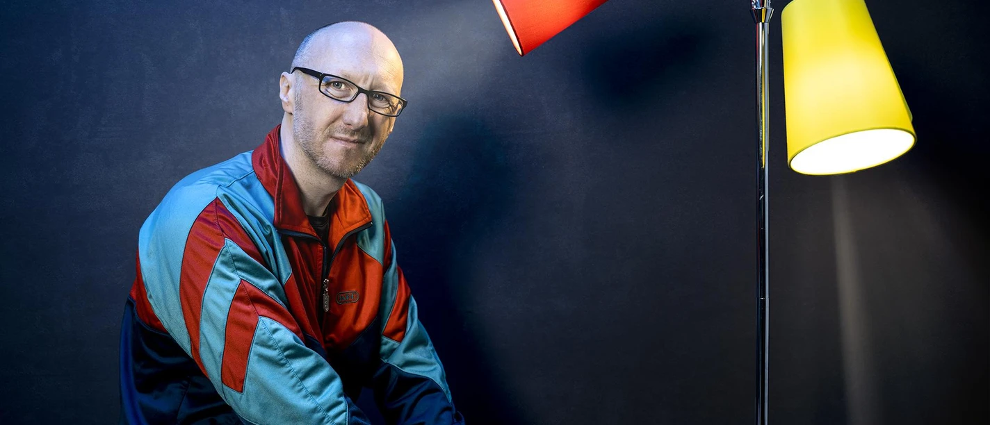 Ralf Senkel mit Brille in bunter Jacke neben roter und gelber Stehlampe