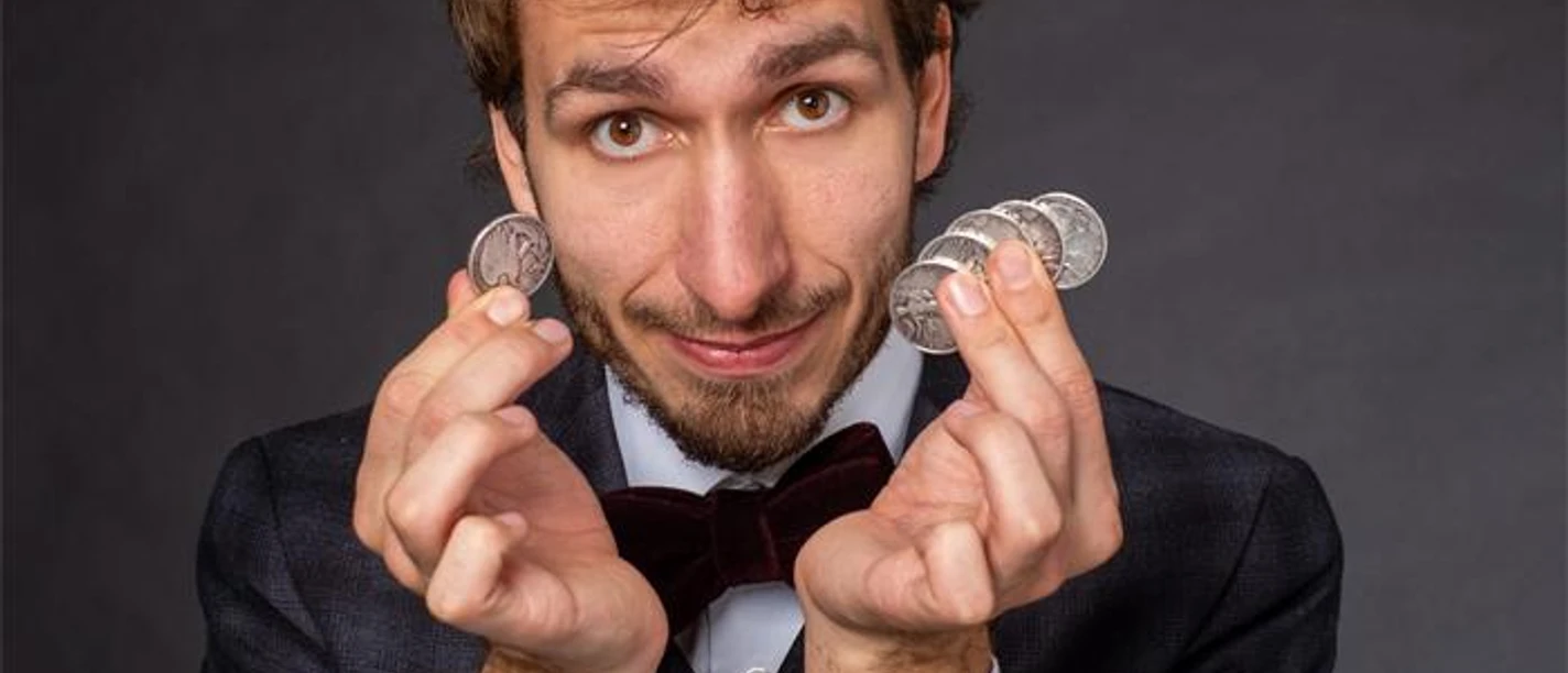 Toby Rudolph im Anzug mit Fliege präsentiert Münzen vor dunklem Hintergrund