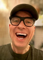 HG Butzko lacht in die Kamera, mit dunkler Brille und Schiebermütze vor warmem Hintergrund.