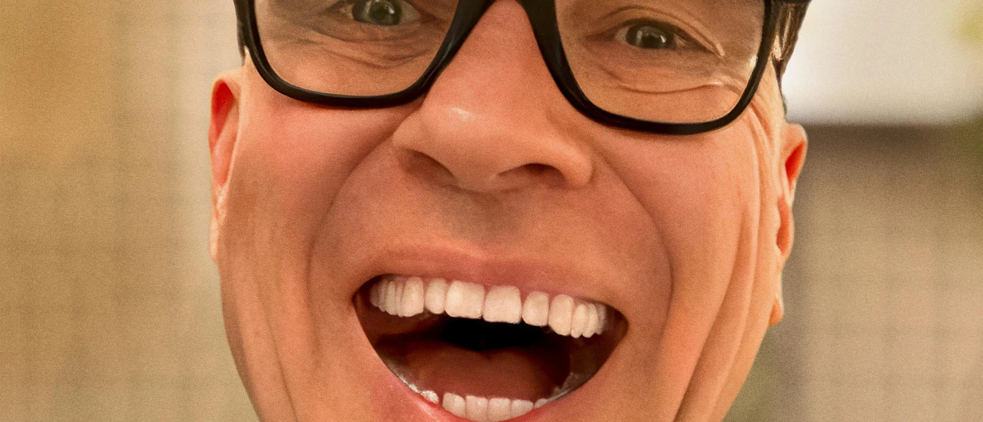 HG Butzko lacht in die Kamera, mit dunkler Brille und Schiebermütze vor warmem Hintergrund.