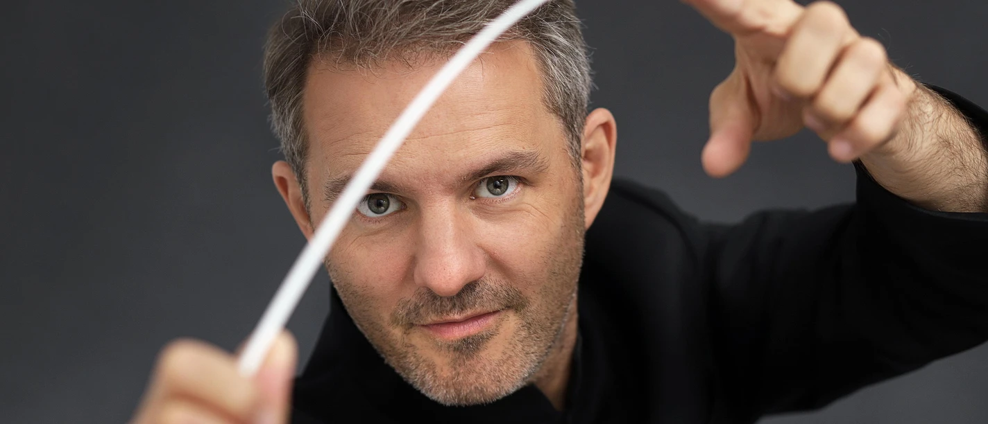 Dirigent Johannes Klumpp mit Taktstock vor dunklem Hintergrund