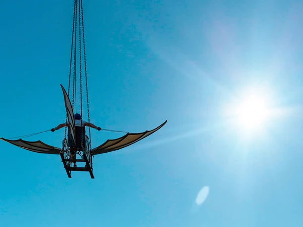 Fliegender Rollstuhl mit Flügeln vor blauem Himmel und strahlender Sonne