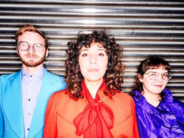 Sheen Trio in colorful outfits in front of a metallic roll-up gate at the Multiphonics Festival