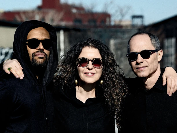 Three musicians wearing sunglasses pose arm in arm against an urban backdrop