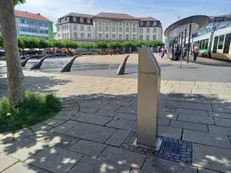 Im Vordergrund befindet sich der Trinkbrunnen. Im Hintergrund ist der Königsplatz sichtbar mit den Wasserspeiern, Gebäuden und zur rechten der Haltestelle auf dem Königsplatz.