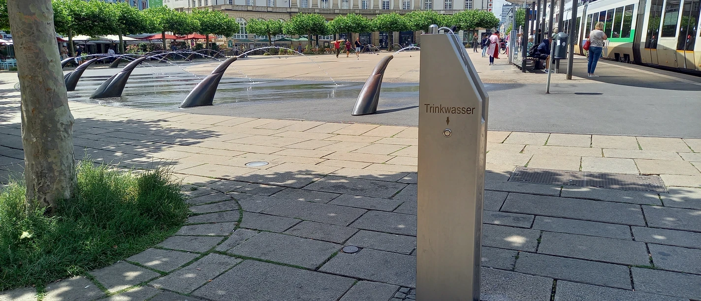 Trinkbrunnen Königsplatz Im Vordergrund befindet sich der Trinkbrunnen. Im Hintergrund ist der Königsplatz sichtbar mit den Wasserspeiern, Gebäuden und zur rechten der Haltestelle auf dem Königsplatz.