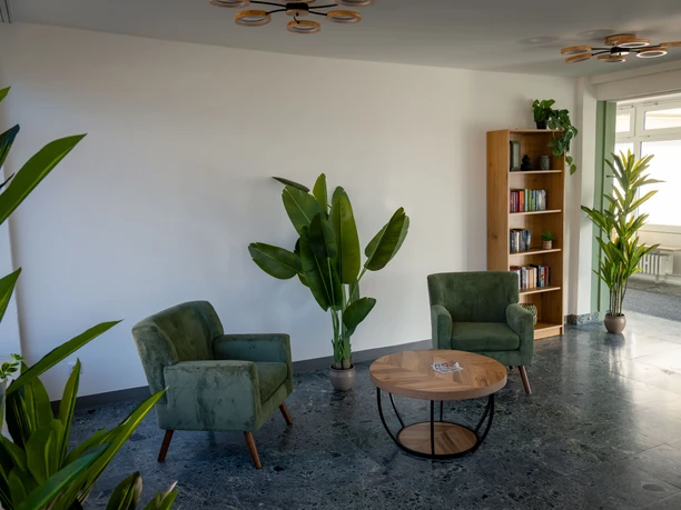 Aufenthaltsraum mit zwei grünen Sesseln, rundem Holztisch, Pflanzen und einem Bücherregal.