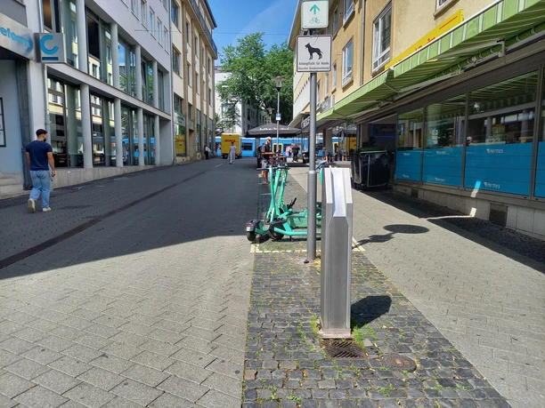 Sie Blicken entlang einer Straße, der Trinkbrunnen befindet sich gleich zu Anfang der Straße. Links und rechts sind Häuser abgebildet.