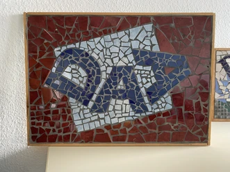 DAA_Mosaik.jpeg Gerahmtes Mosaik mit blauen DAA-Buchstaben auf rotem und weißem Scherbenhintergrund.