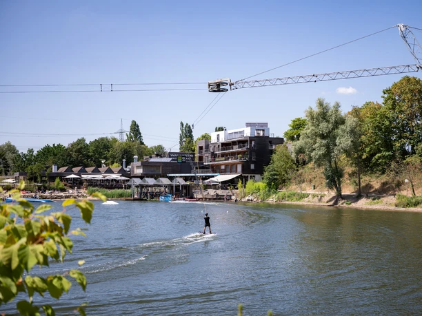 Person beim Wasserski auf dem See der Anlage Langenfeld, mit Seilzug und den Gebäuden der Wasserskianlage am Ufer