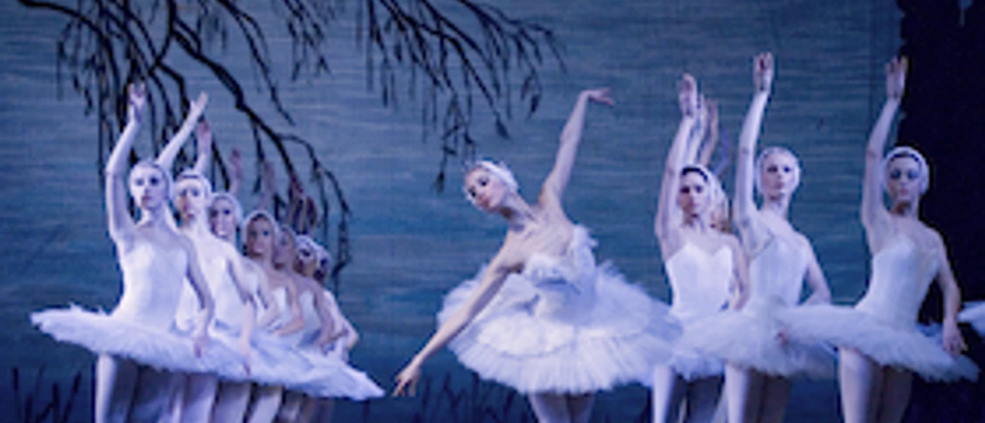 Ballettensemble in weißen Tutus tanzt Schwanensee auf blau beleuchteter Bühne.
