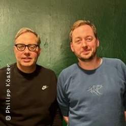 Zwei Männer vor grüner Wand, einer mit Brille und schwarzem Pullover.
