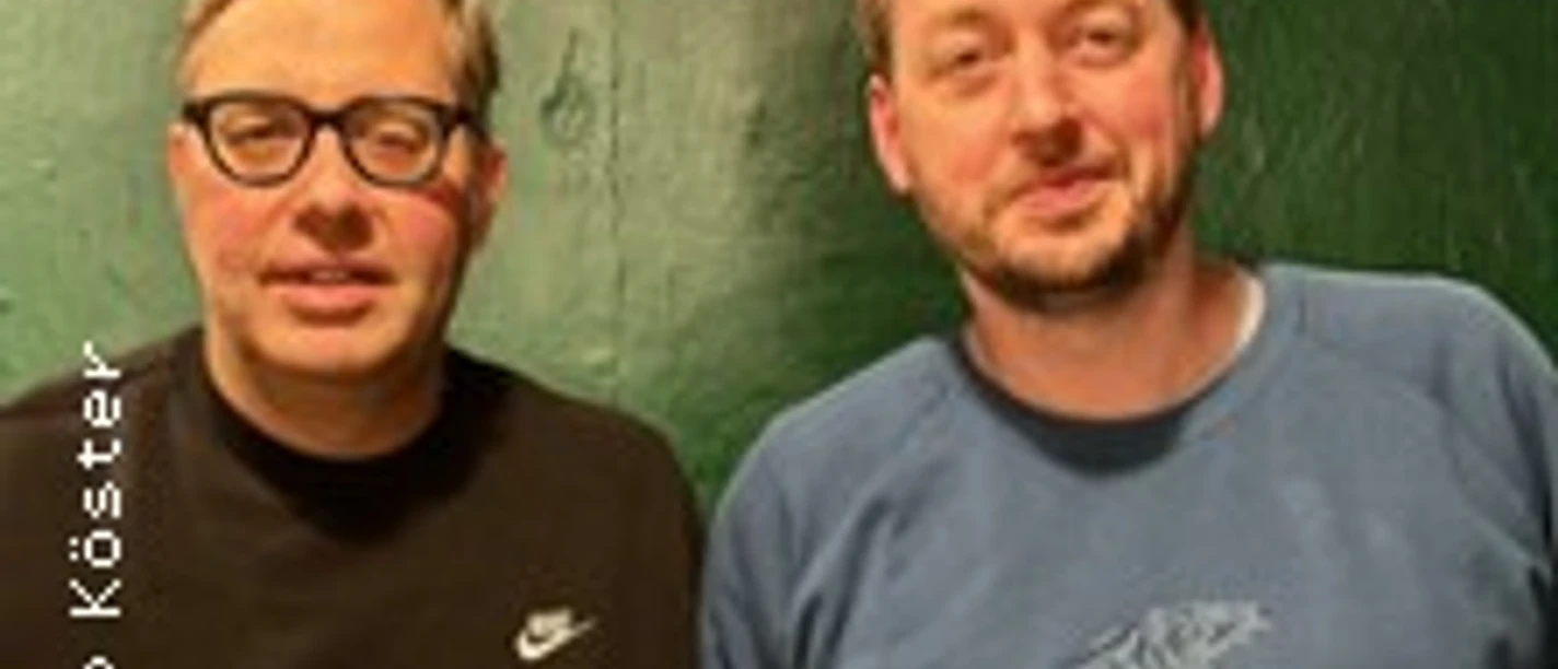 Zwei Männer vor grüner Wand, einer mit Brille und schwarzem Pullover.