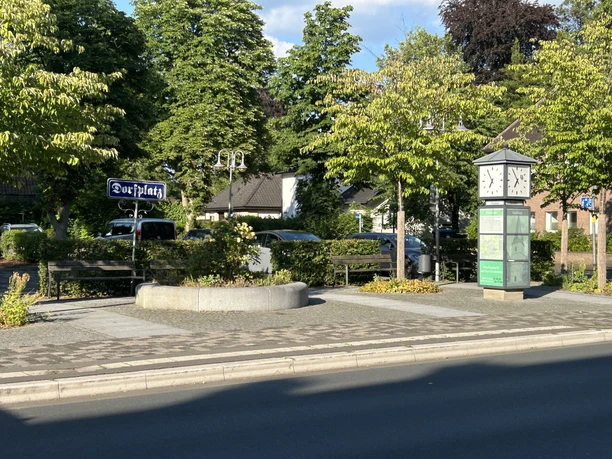 Eselsweg Dorfplatz mit Schild, Uhr, Infotafel und Bänken zwischen grünen Bäumen.
