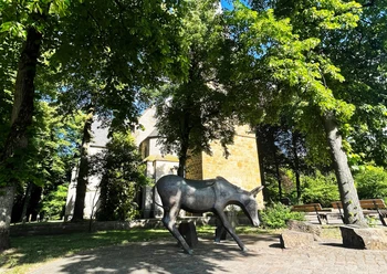 Eselsweg Bronzene Eselstatue auf gepflastertem Platz vor einer Kirche unter Bäumen.