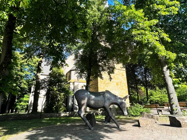 Eselsweg Bronzene Eselstatue auf gepflastertem Platz vor einer Kirche unter Bäumen.