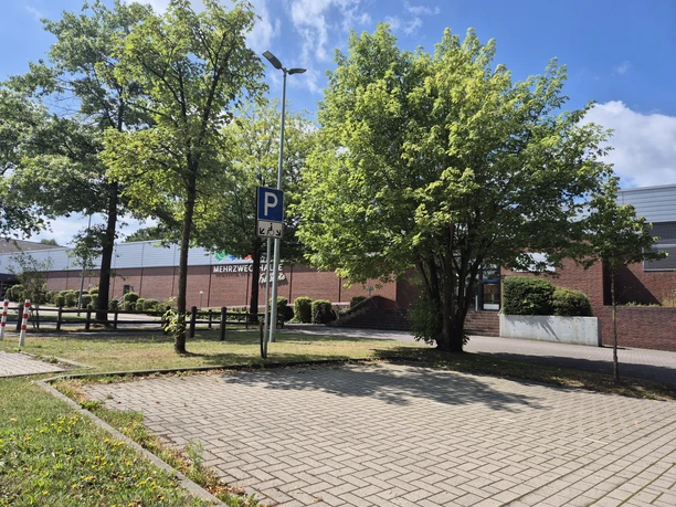 Parkplatz an der Mehrzweckhalle Feldbreite mit P-Schild, Bäumen und blauem Himmel.