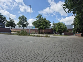 Gepflasterter Parkplatz vor der Mehrzweckhalle Feldbreite, umgeben von Bäumen.