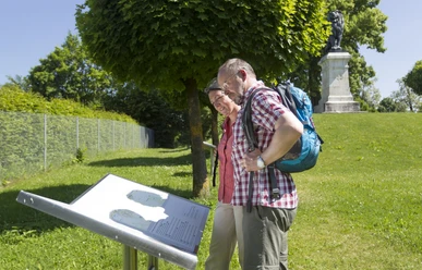 Löwendenkmal in Schierling/Eggmühl Zwei Personen betrachten eine Informationstafel vor dem Löwendenkmal im Grünen.