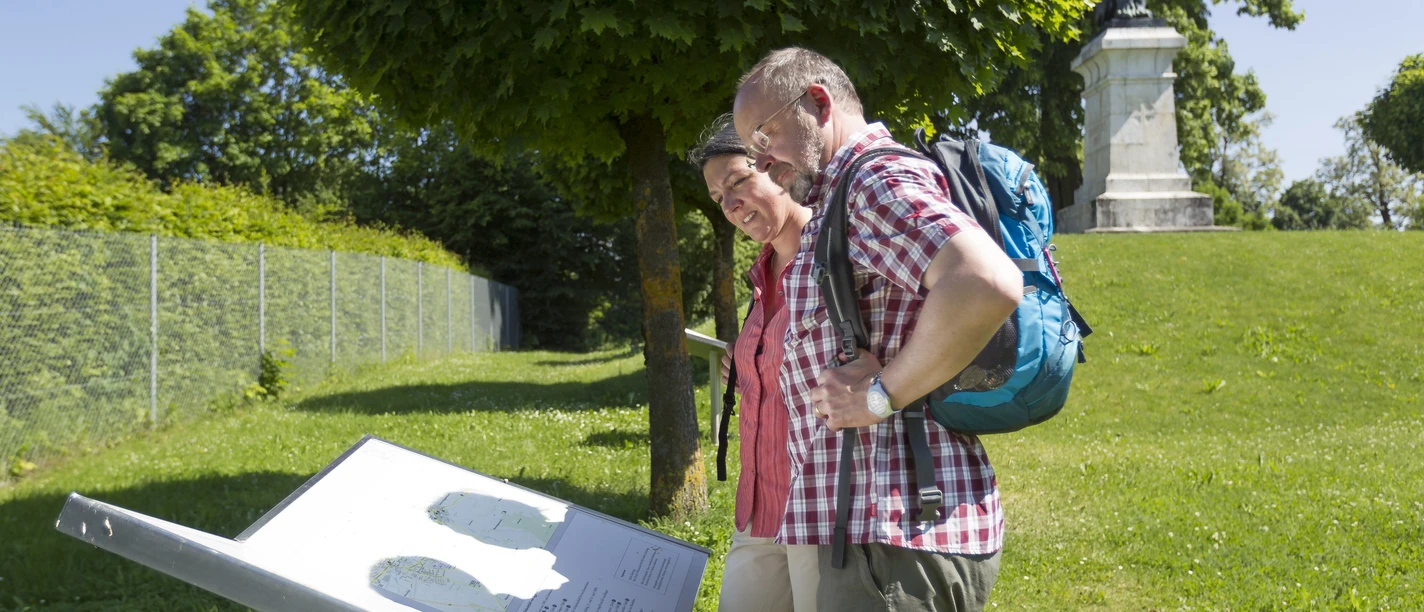 Löwendenkmal in Schierling/Eggmühl Zwei Personen betrachten eine Informationstafel vor dem Löwendenkmal im Grünen.
