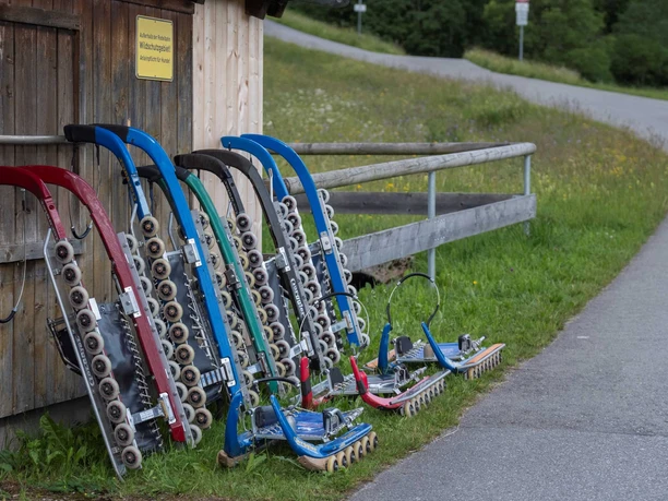 Mehrere Rollenrodel lehnen an einer Holzhütte neben einer asphaltierten Bahn im Grünen