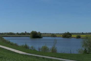 Baggersee in Rade mit Uferweg, grünen Wiesen und Feldern unter blauem Himmel.