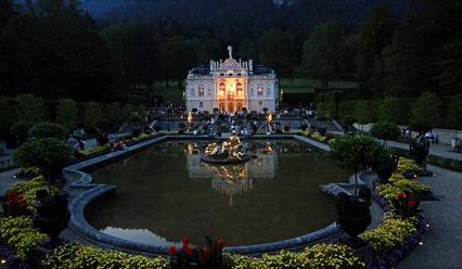 Erleuchtetes Schloss Linderhof bei Nacht mit Wasserbecken, Brunnen und Gästen im Park