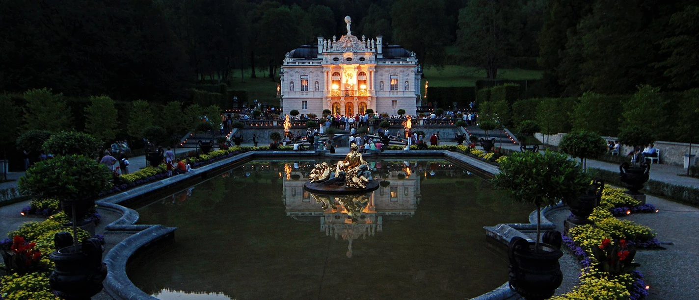 Erleuchtetes Schloss Linderhof bei Nacht mit Wasserbecken, Brunnen und Gästen im Park