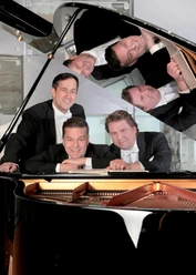 Eine Gruppe von vier Männern in eleganter Kleidung lächelt und posiert um ein Klavier. Ihre Gesichter spiegeln sich im Klavier, was eine interessante Perspektive schafft.