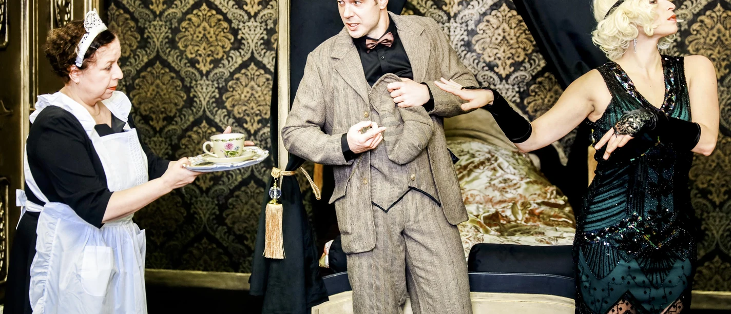 Theaterszene mit Dienstmädchen, elegantem Paar und Teetablett vor goldener Tapete
