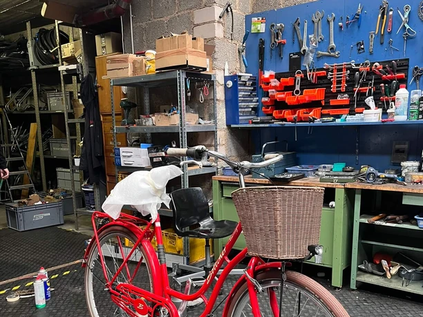 A red bicycle with a basket in a Cologne bike shop, in front of a wall of tools and a workbench