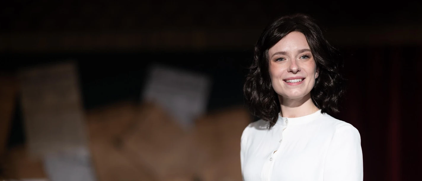 THE ARTIFICIAL SILK GIRL An actress in a white blouse smiles on a theater stage against a dark background