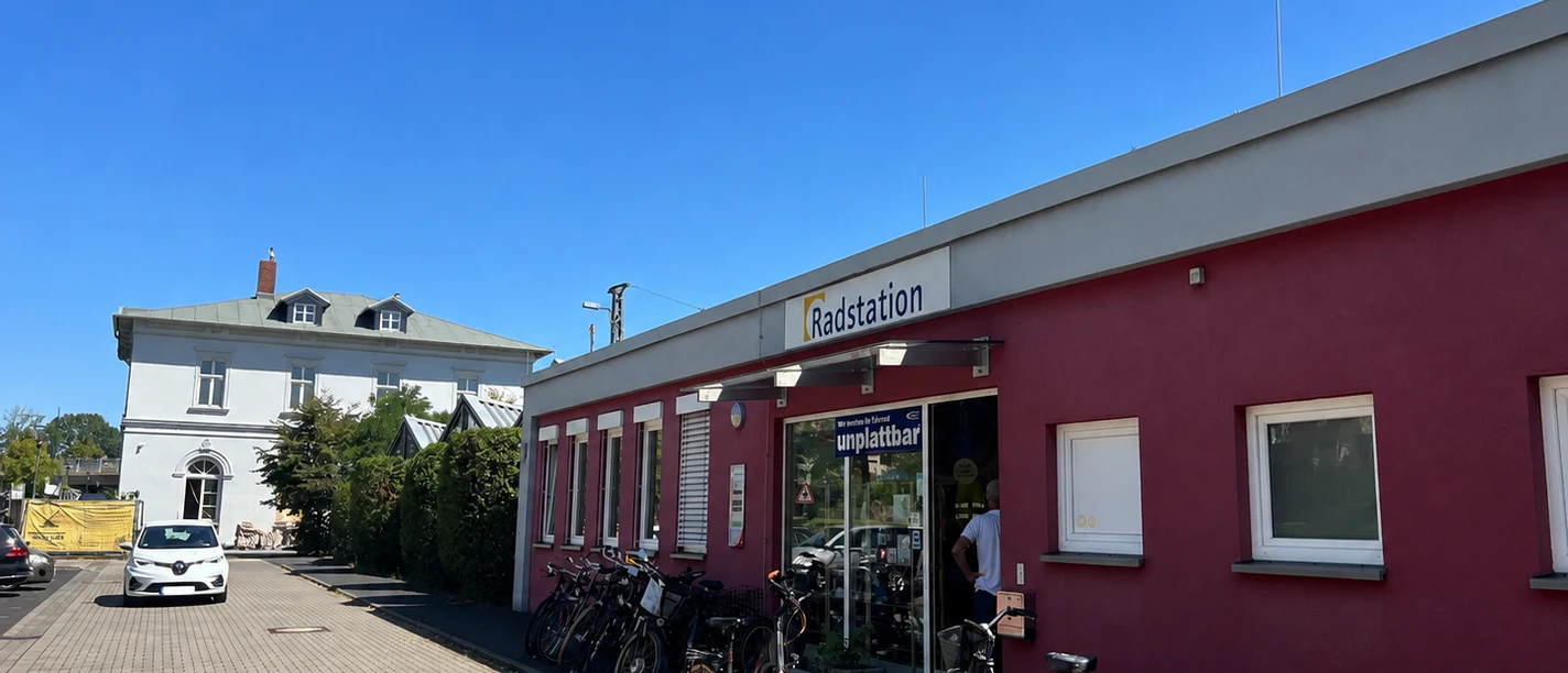 Rotes Gebäude der Radstation mit abgestellten Fahrrädern vor einem weißen Haus bei blauem Himmel.