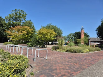 Marktplatz Rastede mit Ziegelpflaster, Fahrradständern, Grünflächen und rotem Turm.