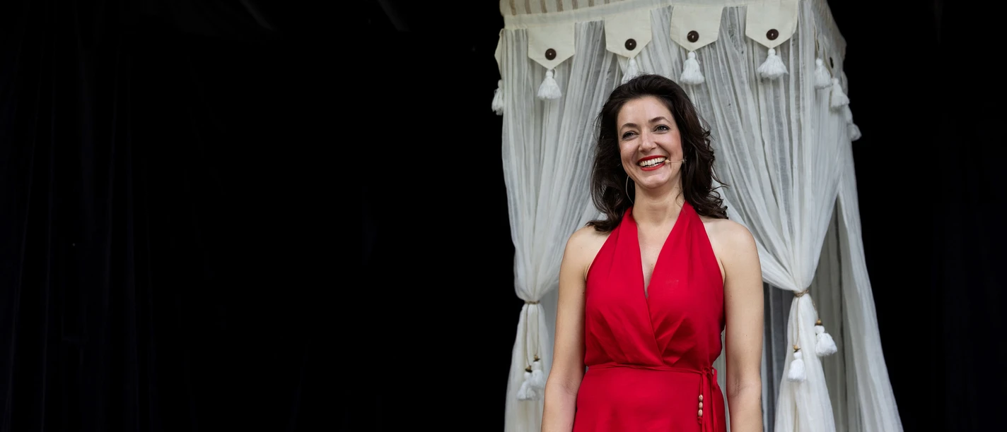 DAS LÄCHELN DER FRAUEN – Komödie von Nicolas Barreau Lachende Schauspielerin im roten Kleid vor weißem Bühnenvorhang