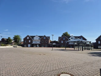 Gepflasterter Dorfplatz Hahn-Lehmden mit Glasunterstand und Backsteinhäusern.