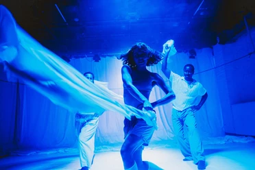 Theater-Performance: TOUCH THE LINE Drei Tänzerinnen und Tänzer schwingen weiße Stoffbahnen in blauem Bühnenlicht.