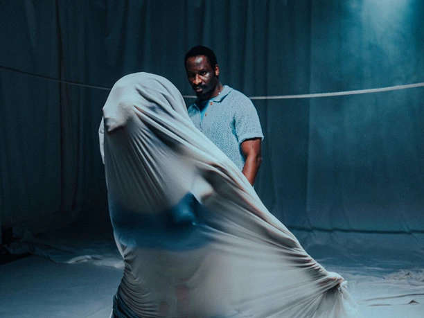Theater-Performance: TOUCH THE LINE Performer auf dunkler Bühne neben einer in weißes Tuch gehüllten Figur