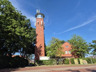 Alter Feuerwehrschlauchturm aus Backstein neben Bäumen und rotem Gebäude unter blauem Himmel.