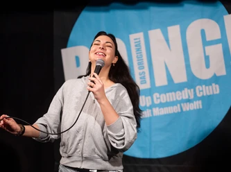 Boing! Comedy Club - Open Mic Extreme A smiling comedienne holding a microphone on stage in front of the BOING! Comedy Club banner