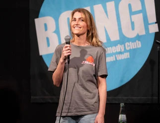 BOING! Comedy Open Mic Köln Ehrenfeld Komikerin mit Mikrofon vor BOING!-Comedy-Club-Logo auf der Bühne