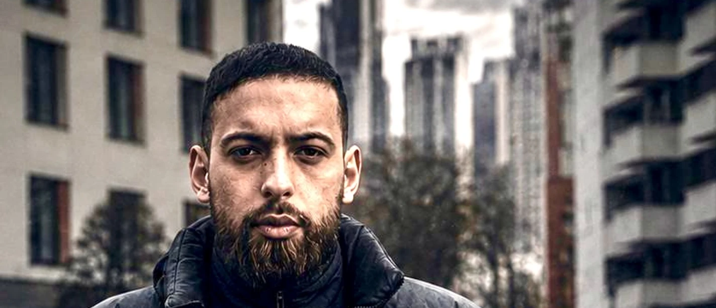 Underground Rapper „Ça va?“ Mann mit Bart in schwarzer Jacke steht vor städtischer Kulisse mit Hochhäusern.