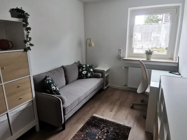 Wohnraum mit grauem Sofa, floralem Kissen, Schreibtisch am Fenster und Holzfußboden.