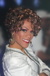 Whitney Houston Tribute Show by Ikenna Amaechi Lächelnde Frau mit lockigem Haar und großen Ohrringen in einem weißen Oberteil.