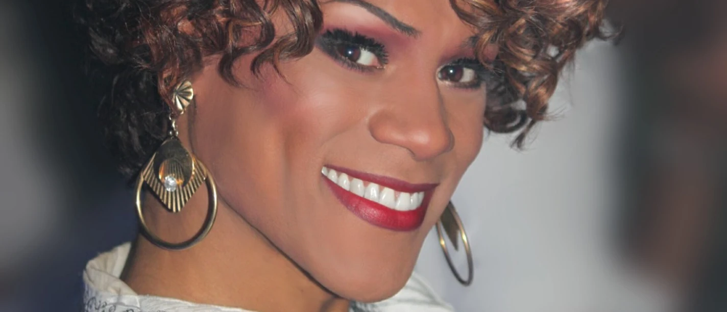 Whitney Houston Tribute Show by Ikenna Amaechi Lächelnde Frau mit lockigem Haar und großen Ohrringen in einem weißen Oberteil.