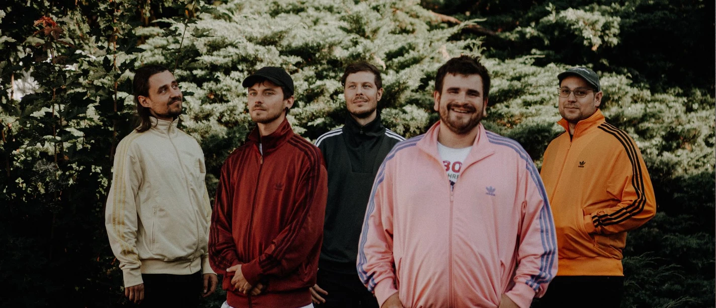 Bandfoto Pizza Hawaii der Film Fünf Männer in bunten Trainingsjacken stehen vor grünen Sträuchern.
