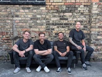 DOMI BADE Collection Four men in black T-shirts are sitting on a sofa in front of a brick wall.