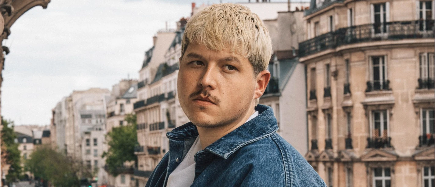 Nio Mann mit blondem Haar und Jeansjacke blickt in einer Stadtlandschaft zur Seite.