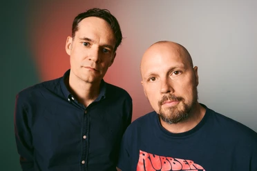 Two men in front of a multicolored background; one is wearing a shirt, the other a T-shirt.